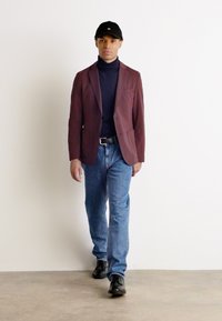 Burgundowa marynarka na granatowym golfie, niebieskie dżinsy i czarne buty. Model nosi czapkę w kolorze czarnym, stojąc na jasnym tle.
