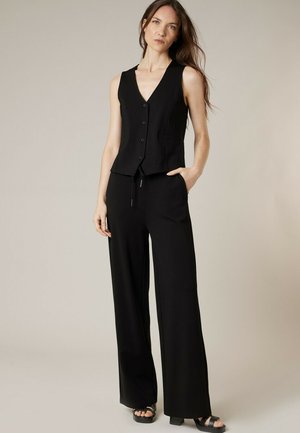 Femme aux longs cheveux bruns portant un gilet sans manches noir boutonné, un pantalon noir à jambes larges et des sandales plateformes noires, debout avec une main dans la poche.