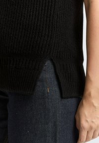 Schwarzer Strickpullover mit geripptem Muster und geteiltem Saum. Trägt man über dunklen Jeans, wodurch die Form und Details des Kleidungsstücks hervorgehoben werden.