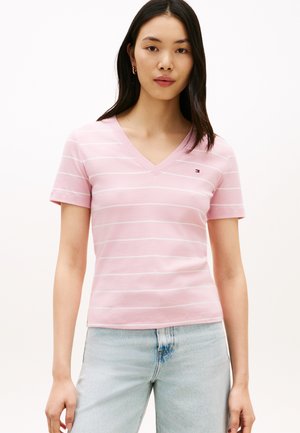 Jonge vrouw met lang donker haar, gekleed in een lichtroze gestreept V-hals T-shirt en lichtblauwe spijkerbroek, staand tegen een effen witte achtergrond.