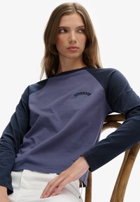 Młoda kobieta o prostych brązowych włosach, ubrana w fioletowo-granatową koszulę z długim rękawem z napisem "Superdry", siedzi na drewnianym krześle, patrząc na bok.