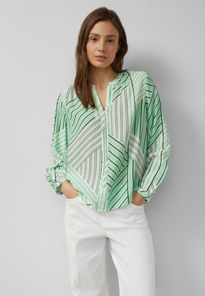 Junge Frau mit langen braunen Haaren trägt eine lockere grün-weiß gestreifte Bluse und weiße Hosen, steht mit den Händen in den Taschen.