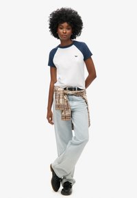 Kortærmet, hvid og marineblå raglan T-shirt, lyseblå brede jeans, ternet skjorte knyttet om livet, og sorte sko med strukturerede såler.