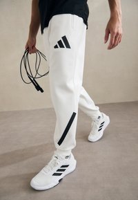 Białe spodnie sportowe z czarnymi akcentami i logo, w połączeniu z białymi butami sportowymi z czarnymi paskami. Model trzyma skakankę.