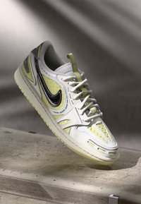 Bijele tenisice s žutim naglasima i teksturiranim panelima. Crni Nike logotip, zaobljenim vrhom i dizajnom s vezicama. Gumeni potplat.