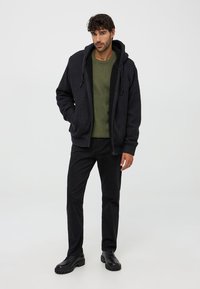 Czarna bluza z zamkiem w teksturowanym materiale z kapturem na sznurku, nałożona na zielony sweter dzianinowy. W połączeniu z czarnymi spodniami i czarnymi butami.