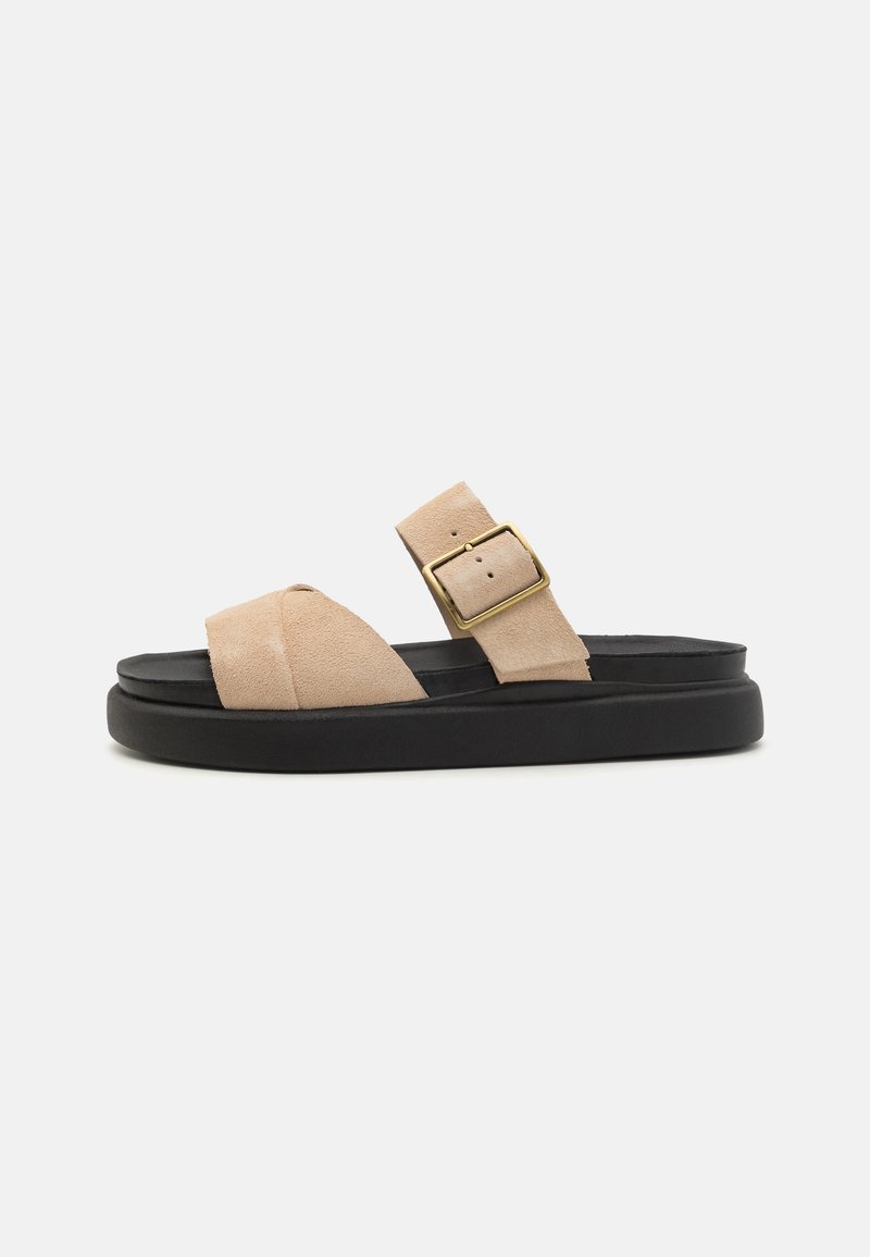Beige suede sandals with a curved strap, gold-tone buckle, and a black platform sole. Flat design suitable for casual wear.