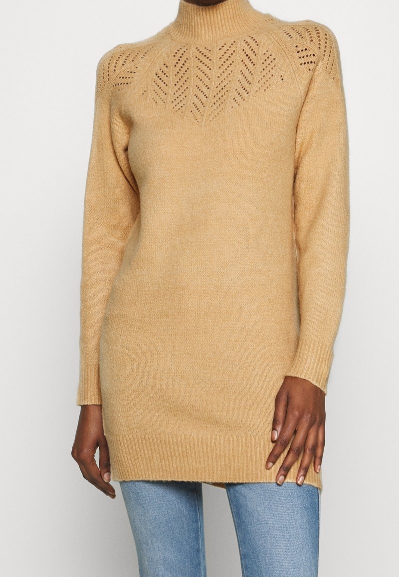Robe-pull beige en maille avec manches longues, ourlet côtelé et motif ajouré décoratif sur le haut de la poitrine et les épaules, portée avec un jean bleu.