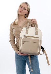 Femme blonde portant un pull beige et un jean, tenant un sac à dos beige et crème avec des poches zippées à l'avant, sur un fond blanc.