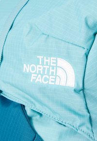 Mėlynas audinio kuprinės modelis su tinklo tekstūra, siuvinėto balto logotipo "THE NORTH FACE" ir kontrastinėmis tamsesnėmis mėlynomis detalėmis.