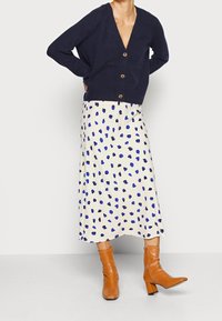 Navy knit cardigan with a V-neck and brown buttons, paired with a cream skirt featuring blue and black abstract spots. Brown square-heeled boots.