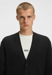 Schwarzer gestrickter Cardigan mit tiefem V-Ausschnitt, ausgestattet mit einem strukturierten Stoff und einem einzigen Knopfverschluss. Über einem hellen Hemd getragen.