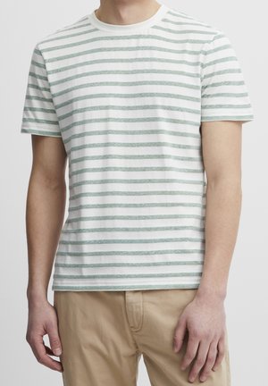 Man draagt een witte T-shirt met korte mouwen en lichtgroene horizontale strepen en beige broek, staand tegen een effen achtergrond.