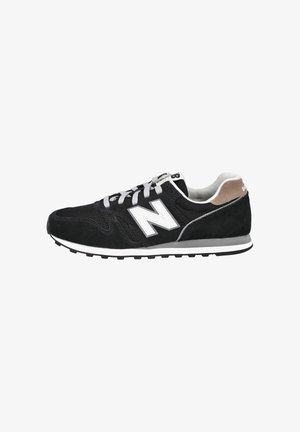 Zapatilla New Balance en blanco y negro con cordones grises, paneles de malla y ante, lengüeta trasera marrón y suela de goma sobre fondo blanco.