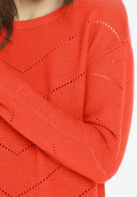 Strikket orange sweater med en løs pasform, der har et mønster af skrå linjer og hulmønsterdetaljer langs ærmerne og kroppen.