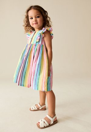 Peutermeisje met krullend haar, gekleed in een mouwloze, meerkleurige gestreepte jurk en witte sandalen, staand op een beige achtergrond.