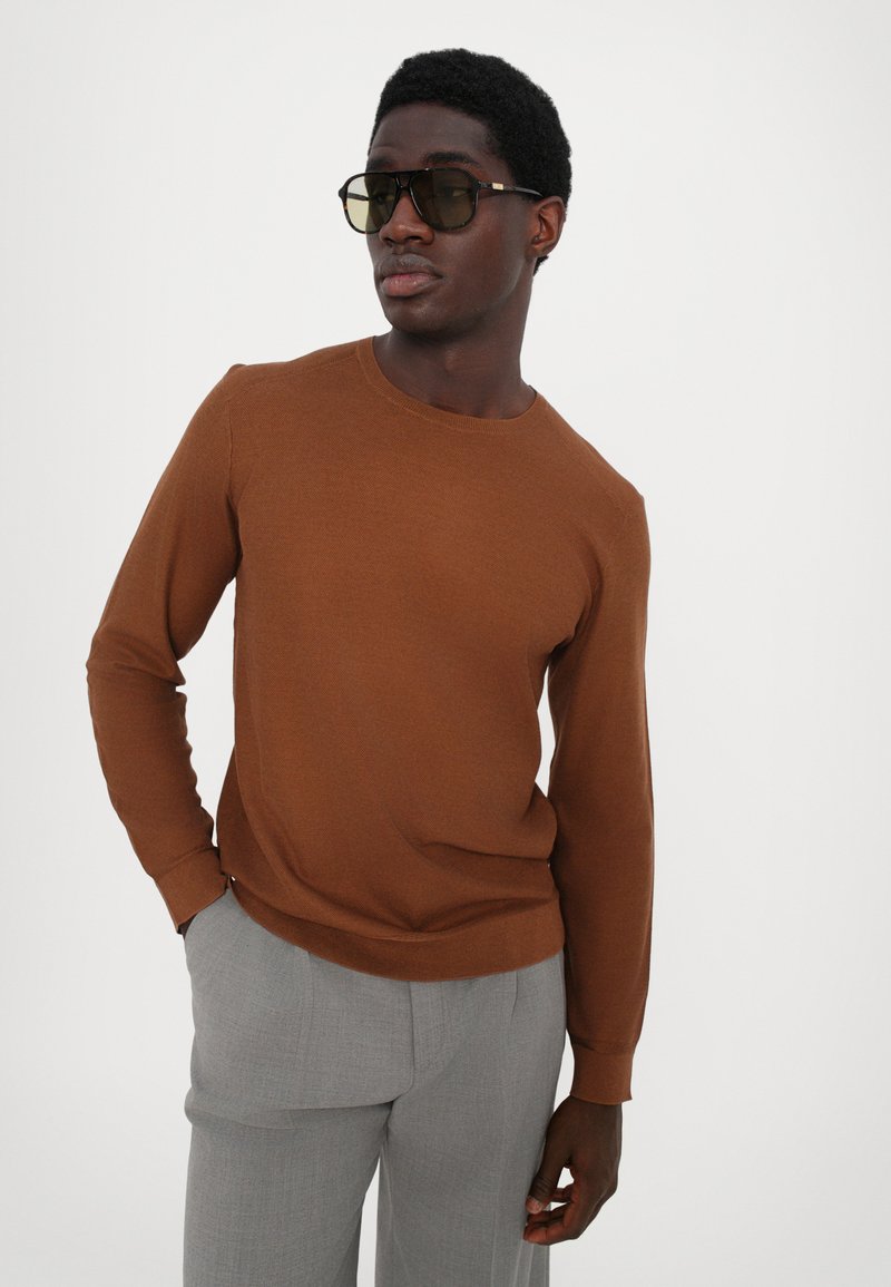 Brun tröja i mjukt stickat tyg med rundad halsringning, matchad med grå skräddarsydda byxor. Modellen bär stora fyrkantiga solglasögon.