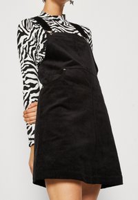 Svart manchesterklänning med justerbara axelband, framfickor och strukturerat tyg. Under bärs en långärmad topp med svartvitt zebramönster.