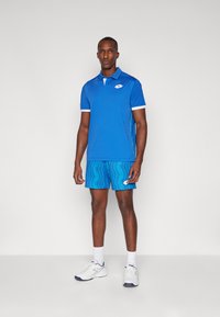 Blue short-sleeve polo shirt with white accents, paired with blue patterned shorts. White athletic shoes complete the sporty look.