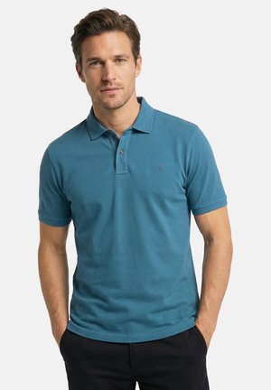 Mann mit kurzen braunen Haaren, der ein türkisfarbenes kurzärmliges Poloshirt und schwarze Hose trägt, mit den Händen in den Taschen vor weißem Hintergrund stehend.