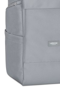 Gray fabric backpack with a flat, rectangular shape, featuring a front pocket, zipper, and a logo patch near the bottom.