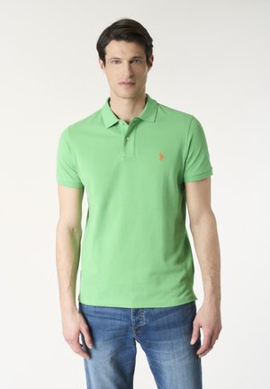 Uomo che indossa una polo verde chiaro con un logo arancione sul petto sinistro e jeans blu, in piedi contro uno sfondo semplice.