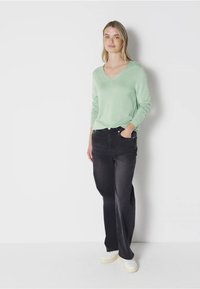 Kobieta stoi z jedną ręką w kieszeni, ubrana w jasnozielony długi rękaw dekolt w serek, czarne szerokie spodnie dżinsowe i białe trampki na białym tle.