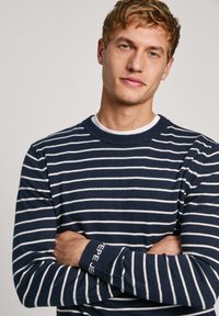 Pletvasta bluza mornarsko plave boje s bijelim prugama, okrugli izrez i rebrasti manžeti. Sadrži logo na manžeti, izrađena od mekog, teksturiranog materijala.