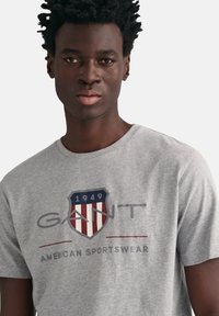 Grijze katoenen T-shirt met een schildlogo met rode, witte en blauwe strepen, en de tekst "GANT" boven "AMERICAN SPORTSWEAR."