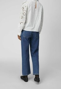 Person som bär en vit ögla-spetsblus, raka blå jeans och svarta loafers, stående mot en enkel grå bakgrund.