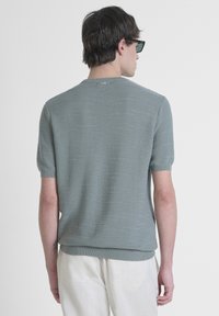 Suéter de punto de manga corta y cuello redondo en verde claro, con un patrón texturizado y dobladillo acanalado. Combinado con pantalones de color claro.