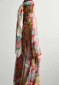 Langes, fließendes Kleid aus leichtem Stoff mit einem lebhaften Blumenmuster in Rot, Grün, Gelb und Blau. Begleitet von einem gemusterten Kopftuch.