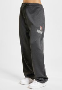 Grå joggebukser med elastisk midjebånd og snøring, med en logo på venstre lår. Kombinert med hvite joggesko.