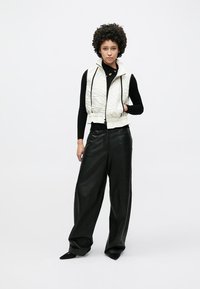 Gesteppte weiße ärmellose Weste mit Frontreißverschluss; strukturierte Oberfläche; kombiniert mit hoch taillierten schwarzen Lederhosen mit weitem Bein. Mit minimalen Akzenten gestaltet.