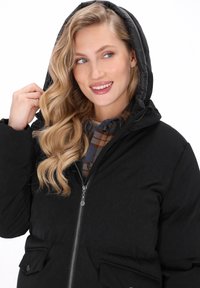 Blonde vrouw met golvend haar lacht terwijl ze de capuchon van een zwarte pufferjack over een bruin en blauw geruit shirt aanpast.