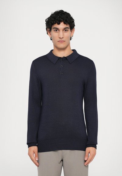 Polo de manga larga azul marino con cuello tipo camisa, tres botones, puños acanalados y una textura suave, combinado con pantalones de color claro.