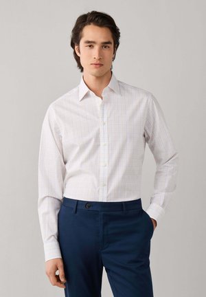 Junger Mann mit dunklen Haaren, der ein weißes Hemd mit feinem Karomuster und dunkelblaue Hose trägt, steht mit einer Hand in der Tasche.