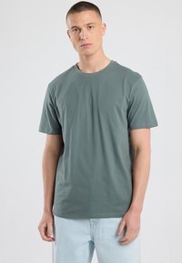 Short-sleeve, crew-neck t-shirt in olive green cotton, featuring a relaxed fit and smooth texture. Worn with light blue jeans.