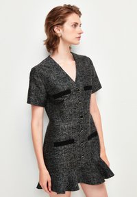 Mustat ja harmaat tweed-mekko lyhyillä hihoilla, V-aukkolla, etunapeilla ja kahdella mustalla taskupaneelilla. Hameosa on rypytetty.