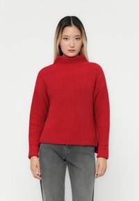 Roter, gestrickter Rollkragenpullover mit strukturiertem Muster, überschnittenen Schultern und gerippten Bündchen. Getragen mit grauen, geraden Jeans.