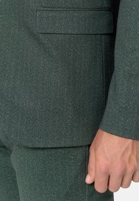 Zeleni strukturalni herringbone odijelo od tkanine s teksturiranim završetkom, s izrezanom reverom, jednim džepom i prilagođenim krojem. Prikaz detalja ruke izbliza.