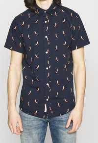 Camisa de manga corta en azul marino con un patrón de chiles naranjas, cuello abotonado y botones blancos. Hecha de tela ligera de algodón.