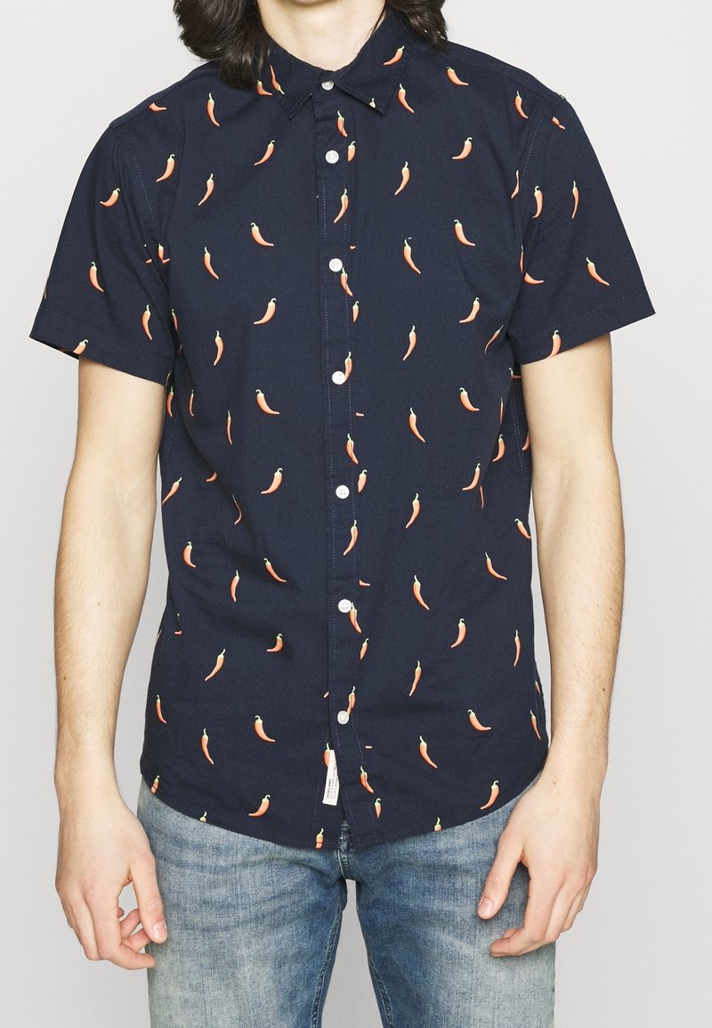 Námořnicky modrá košile s krátkými rukávy, zdobená vzorem oranžových chilli papriček, s límečkem na knoflíky a bílými knoflíky. Vyrobeno z lehké bavlněné látky.