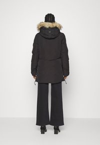 En person står med ryggen mot kameran, iklädd en svart parkas med en huva i päls och vida svarta byxor.