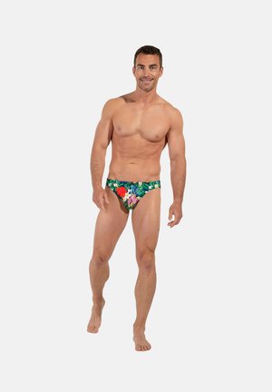 Uśmiechnięty umięśniony mężczyzna stojący boso, ubrany w zielone, kwiatowe kąpielówki, na jednolitym jasnoszarym tle.