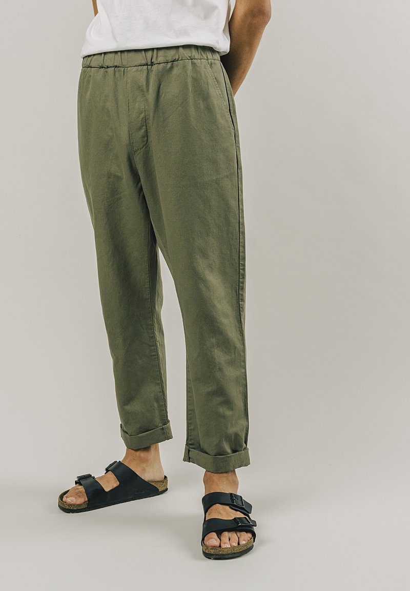 Olivově zelené, lehké zúžené kalhoty s elastickým pasem a bočními kapsami, doplněné černými sandály na dvou popruzích na bosých nohách.