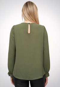 Olivenfarbener Langarmtop mit einem Keyhole-Rücken, strukturiertem Stoff, puffigen Ärmeln und abgerundetem Saum. Glatte Nähte entlang der Kanten.