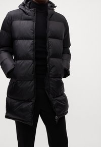 Homme portant une veste matelassée noire à capuche sur un col roulé noir, les mains dans les poches de la veste, sur un fond uni.