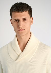 Cremefarbener, gerippter Pullover mit weitem Schalkragen. Der Stoff hat eine glatte Textur und ein tailliertes Design, das den Ausschnitt betont.