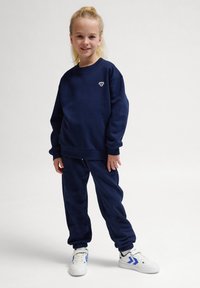 Mörkblå sweatshirt och joggingbyxor gjorda av mjukt tyg, med en liten logotyp på bröstet. Avslutas med vita sneakers med blå detaljer.